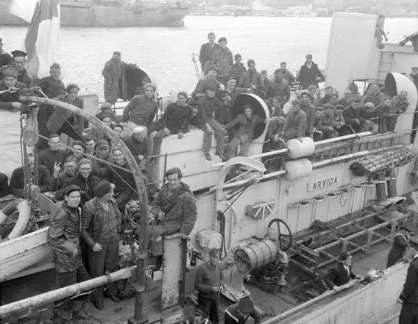 Black and white photograph. The top deck of a ship is crowded with men, sitting and standing on every available surface.